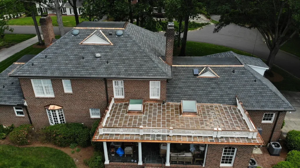 Aerial view of a multi-story brick house featuring a GAF Slateline roof replacement with copper accents and a flat patio roof with two skylights and a white railing, maintained by Charlotte roofing contractors.