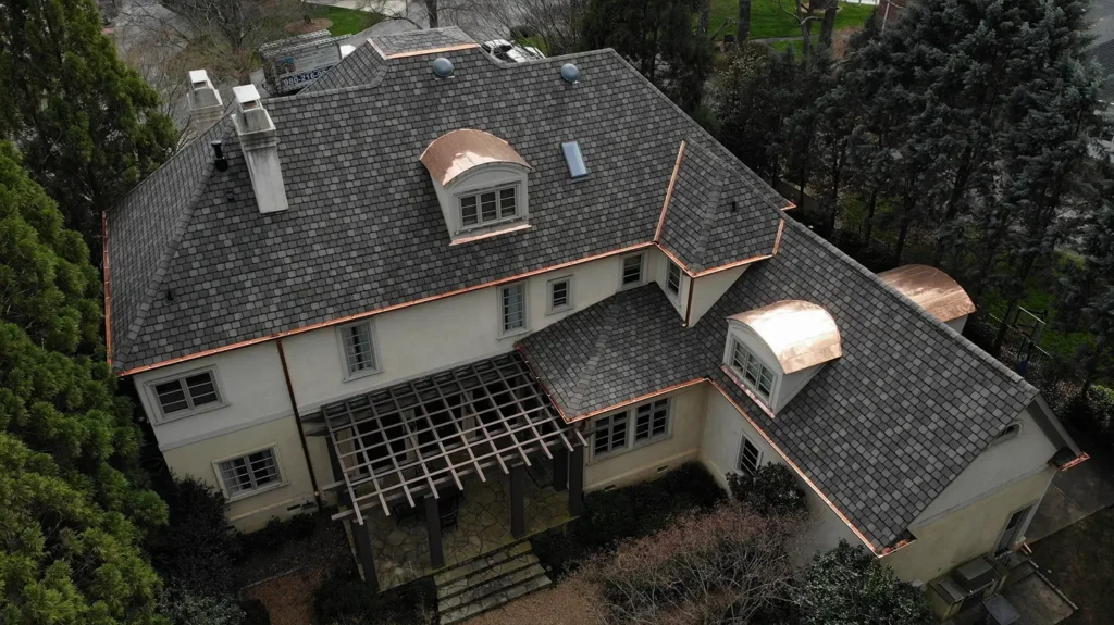 Aerial view of a two-story house featuring a dark slate roof with decorative copper flashing and matching arched copper roofing over dormer windows.