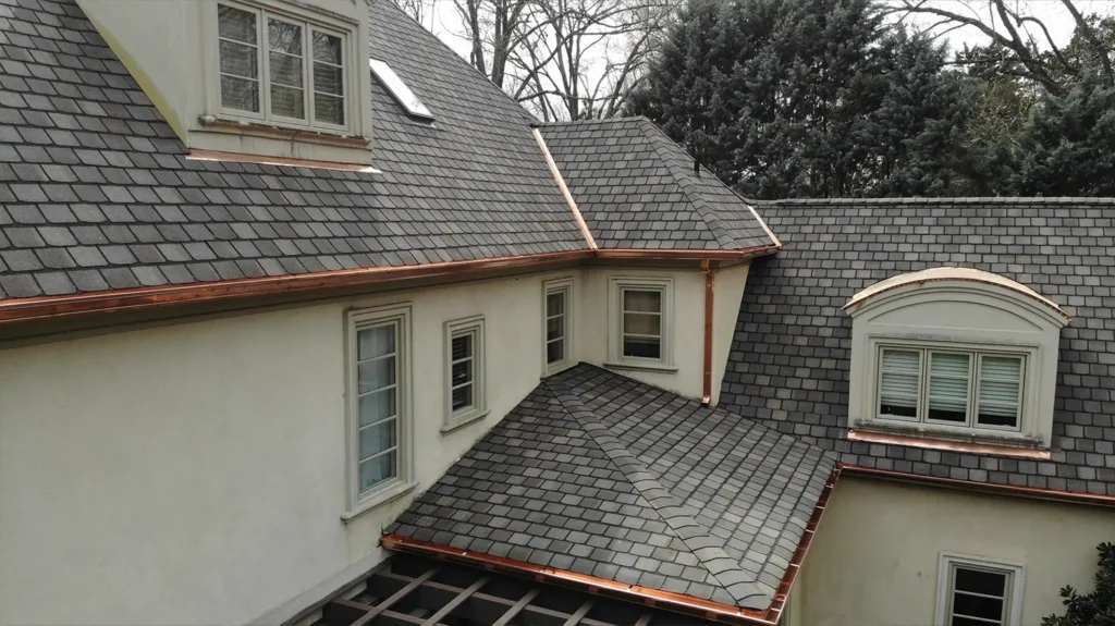An aerial view of a highland slate roof with copper barrel dormers and flashing on a house with light-colored walls.