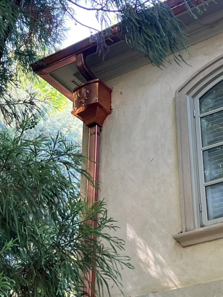 Copper rain gutter downspout with a decorative fleur-de-lis box on a stucco house featuring a Highland slate roof.