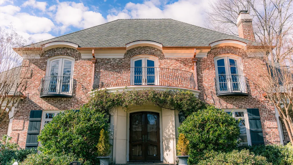 A two-story brick house in Greensboro, NC, featuring three arched windows with small iron balconies on the second floor and a front entrance framed by climbing vines beneath a custom copper roof.
