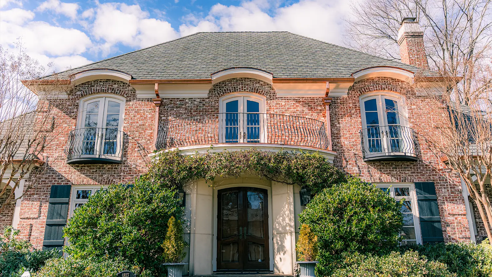 A two-story brick house in Greensboro, NC, featuring three arched windows with small iron balconies on the second floor and a front entrance framed by climbing vines beneath a custom copper roof.