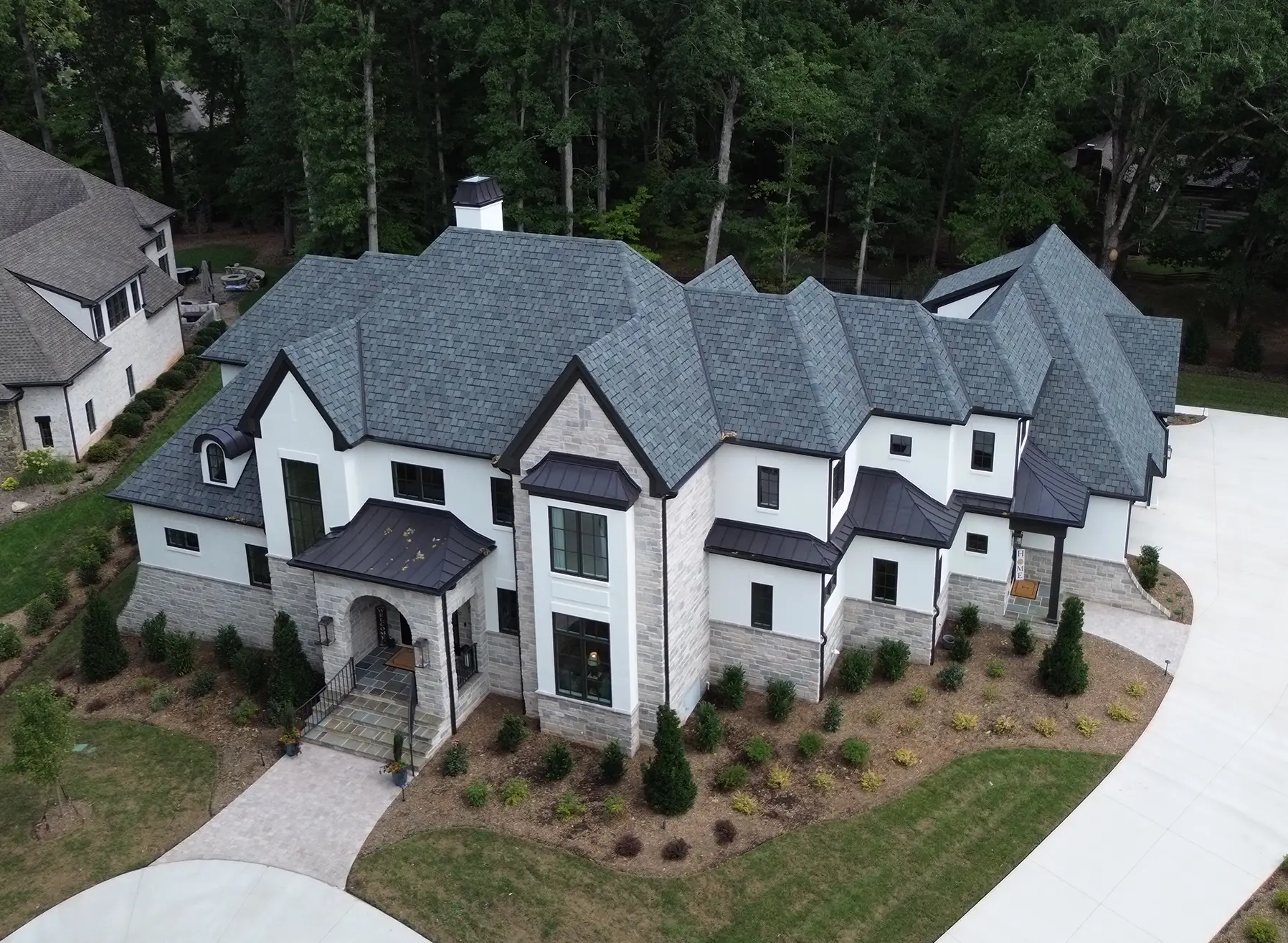 Aerial view of a large, modern two-story house with CertainTeed Belmont shingles and dark bronze metal roofing, featuring white walls and stone accents, surrounded by a landscaped yard and forest.