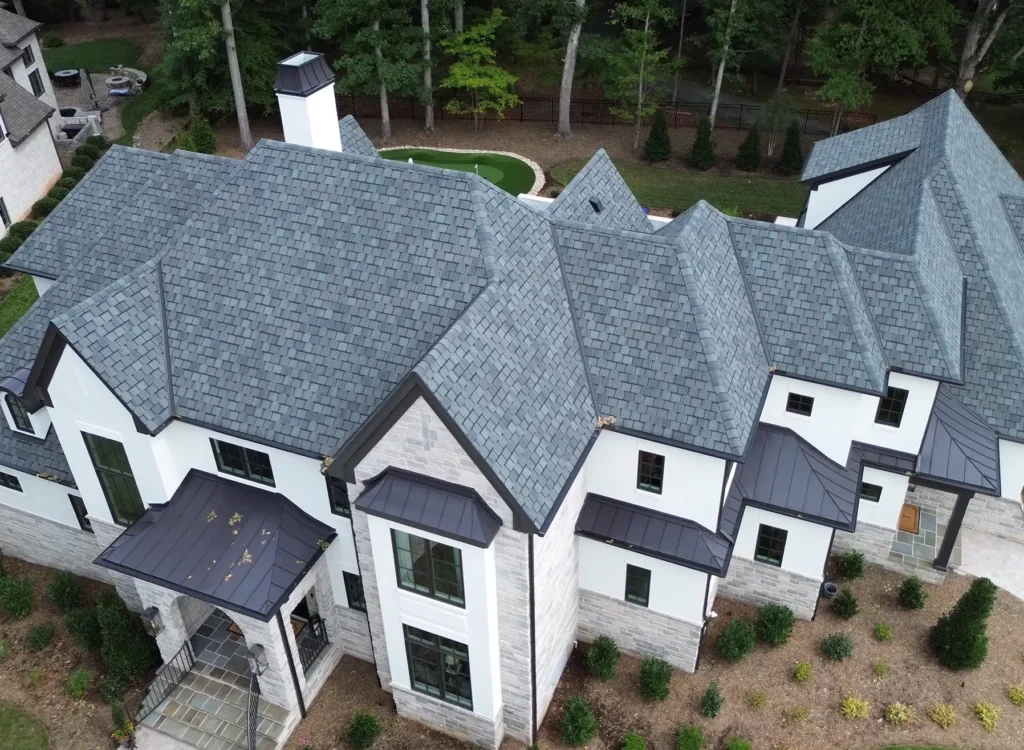 Aerial view of a large, modern house with CertainTeed Belmont shingles, white exterior walls, black trim, and surrounding landscaping, nestled near trees.