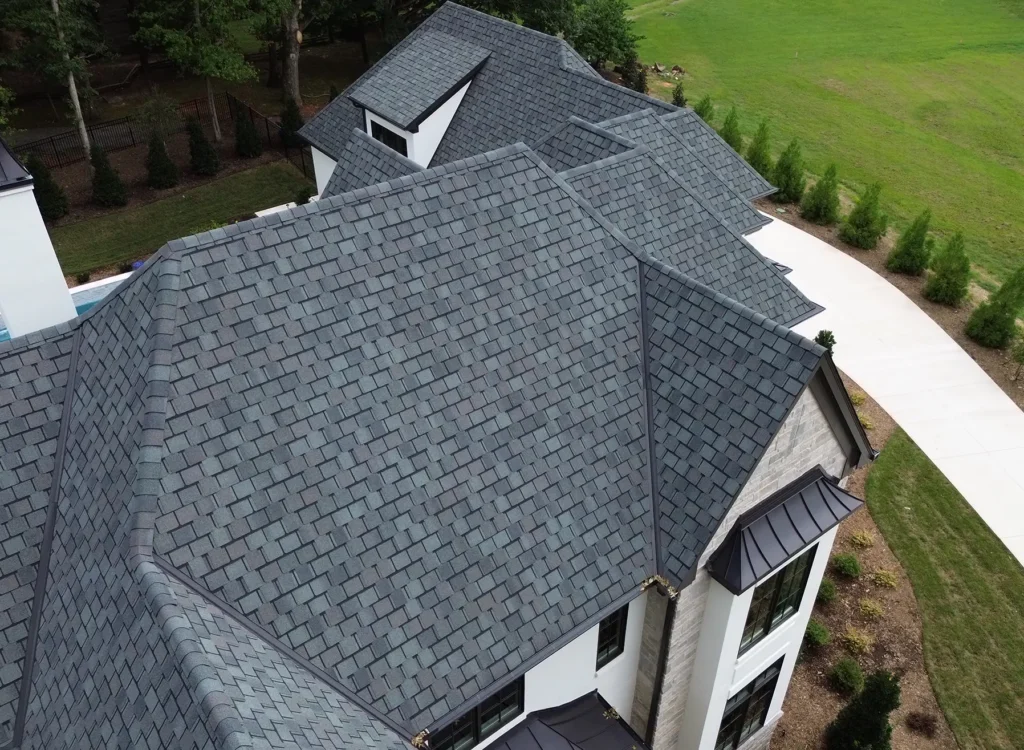 Aerial view of a large house with CertainTeed Belmont shingles on a gray roof, white exterior walls, black window trim, and a driveway leading to a green lawn and trees in the background.