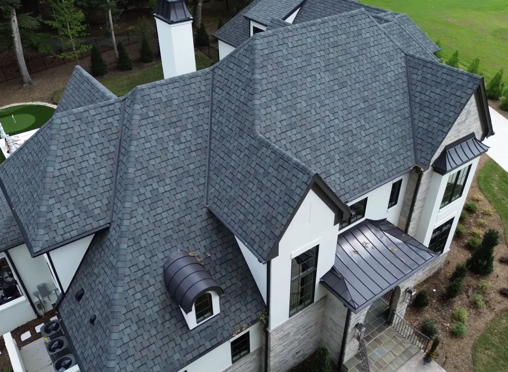 Aerial view of a large modern house with CertainTeed Belmont shingles, white walls, large windows, a dark bronze metal porch roof, and a small dormer. The yard features a putting green and landscaped greenery.