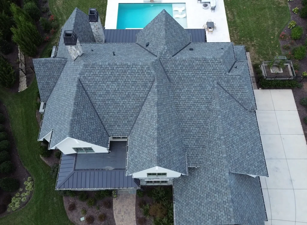 Aerial view of a large house with a complex CertainTeed Belmont shingles roof, surrounded by landscaped greenery, a swimming pool in the backyard, and a concrete driveway on the right side.