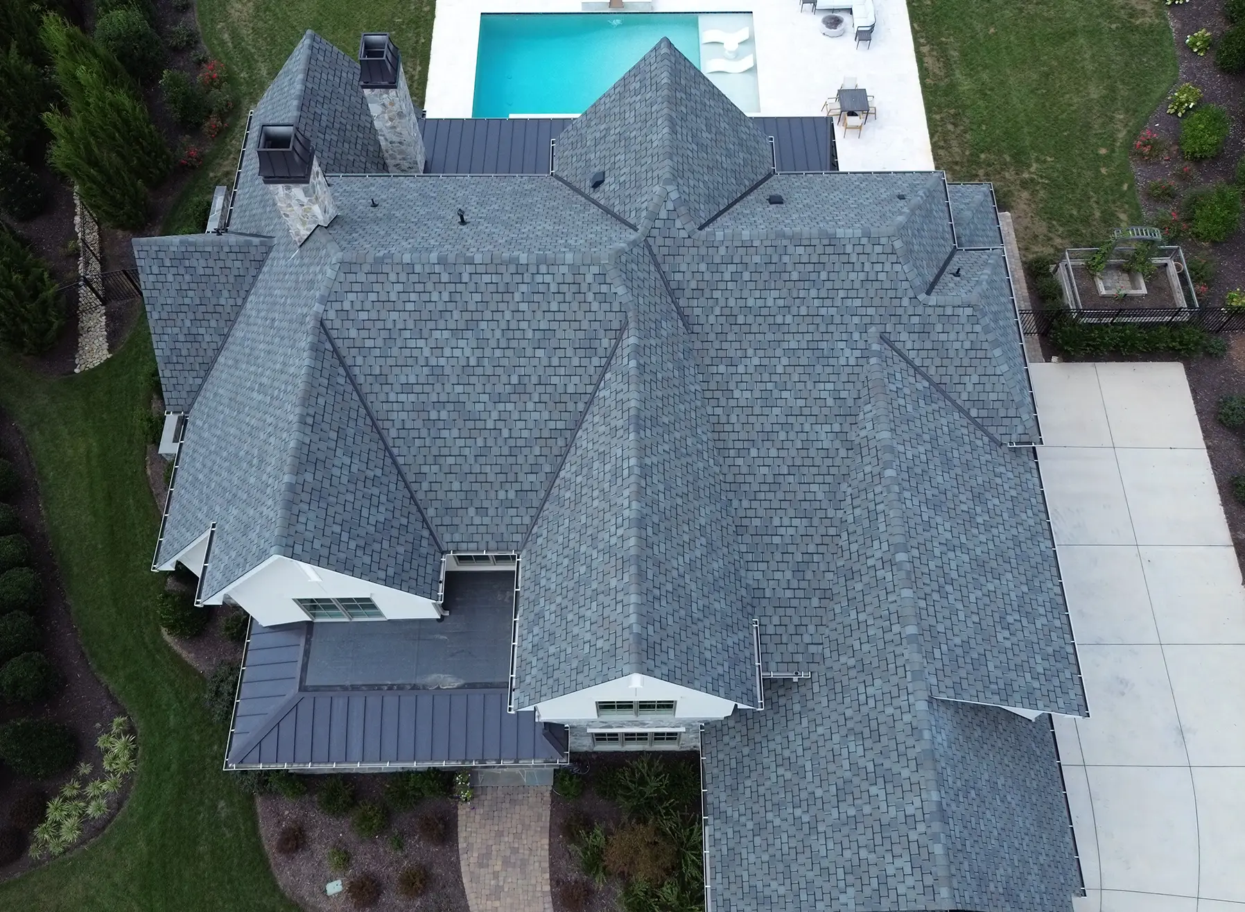 Aerial view of a large house with a complex CertainTeed Belmont shingles roof, surrounded by landscaped greenery, a swimming pool in the backyard, and a concrete driveway on the right side.