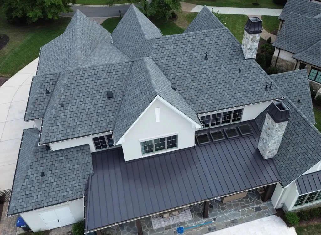 Aerial view of a large house with CertainTeed Belmont shingles, gray shingle roofing, standing seam metal awning, stone chimney, and multiple peaks, surrounded by greenery and paved walkways.