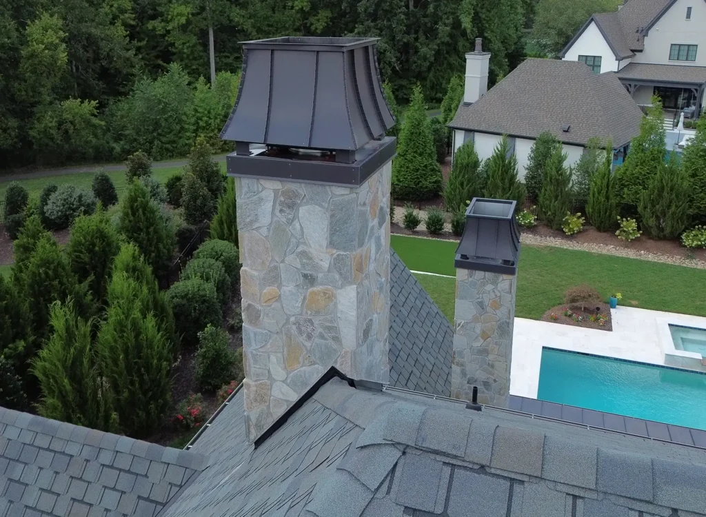 Stone chimneys with metal caps on a house roof featuring CertainTeed Belmont shingles overlook a landscaped yard with a pool, lawn, and trees. Other houses and dense woods are in the background.