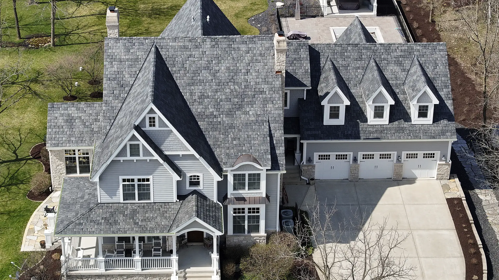 Aerial view of a gray-sided suburban house with a multi-gabled composite roofing style, a three-car garage, and a front porch.