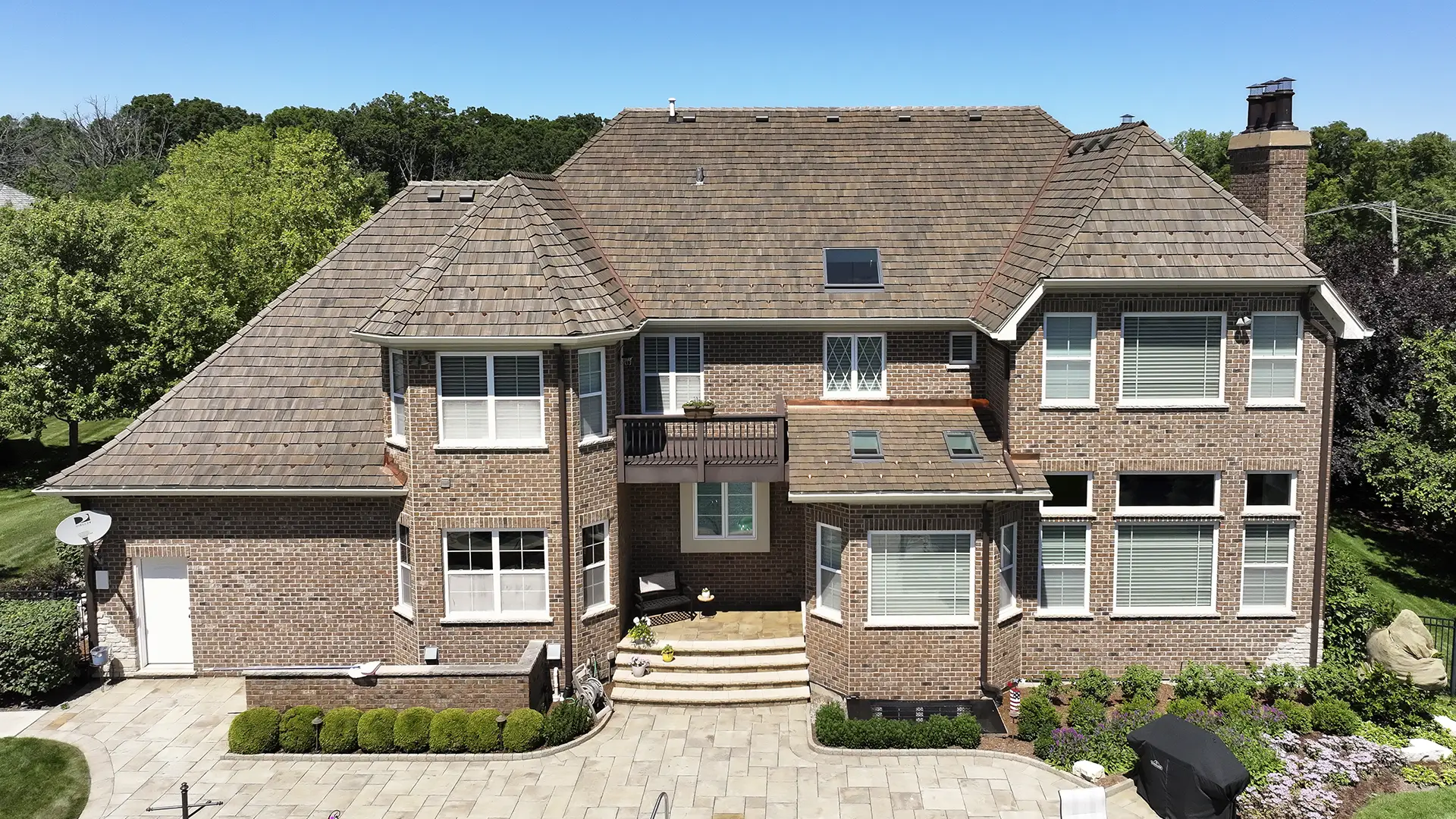 Aerial view of the rear of a two-story brick house with a Brava composite roofing system, a small balcony, and a stone patio.