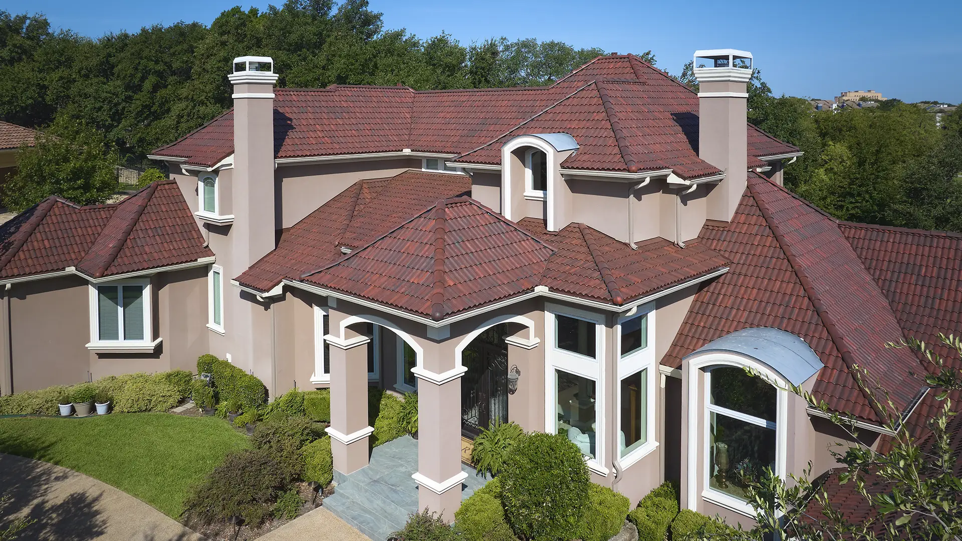 Aerial view of a two-story tan house with a multi-pitched red Brava composite roof and two large chimneys.