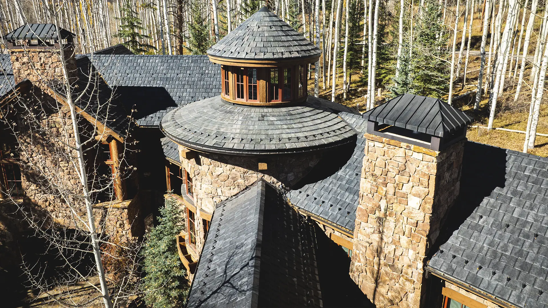 High-angle view of a stone house with a circular turret and Brava composite roofing, nestled in a forest of aspen trees.